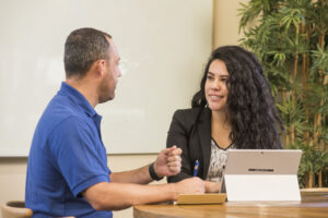 Men wearing a blue t-shirt is talking to a women with long curl brown hair wearing a black suit and white shirt. He is holding a pen and she has her computer in front of her.
