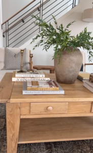 A wood coffee table with a ceramic vase with green plants and four books.