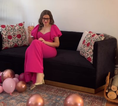 Women in glasses wearing a pink long dress is holding a wine glass seating on a brown couch. The floor is full of pink balloons.