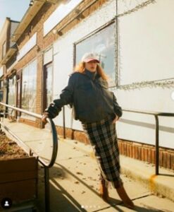 Women wearing a hat standing in a sidewalk wearing a puffy green jacket and a plaid long skirt. 