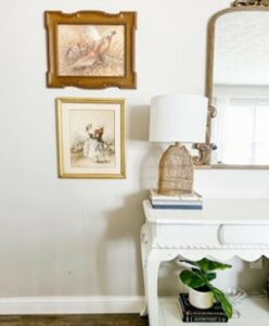 A white console table with a gold mirror and a lamp and two picture frames.