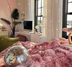 Bedroom picture with fluffy pink comforter and a disco ball on top. A white desk with a green chair next to a plant.