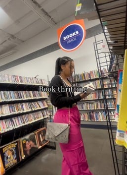 Women wearing pink pants and a black top standing in front of a bookshelf holding a book