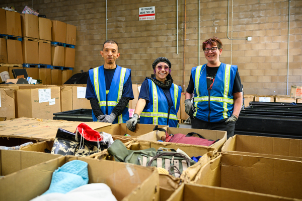 Three people standing behind boxes wearing a blue vest.