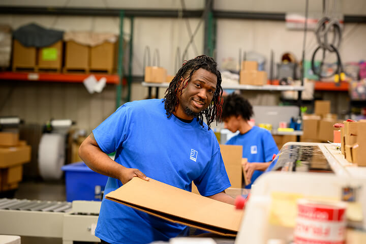 Worker in a blue shirt holds a cardboard sheet in a busy warehouse