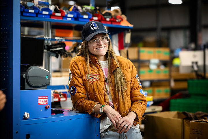 Smiling woman wearing an orange bomber jacket and cap stands at a blue workstation in a busy workshop.