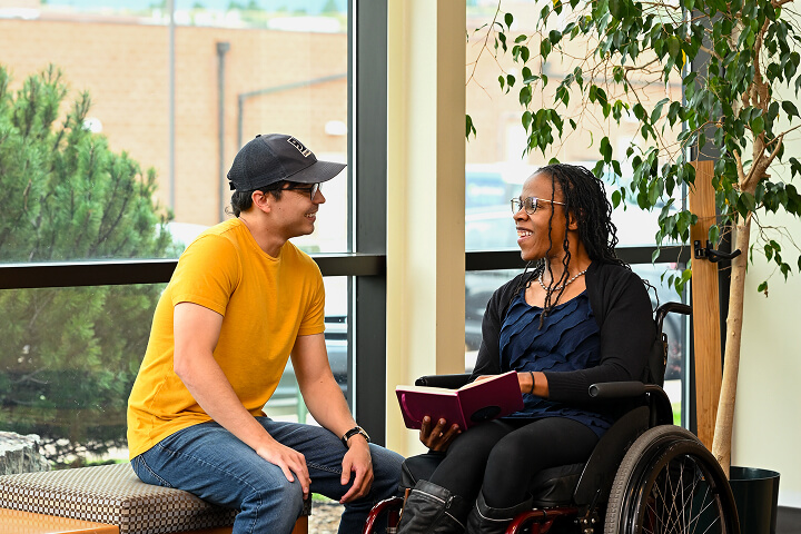 Young man in a yellow t-shirt and cap chats with a woman in a wheelchair indoors, holding a red notebook.