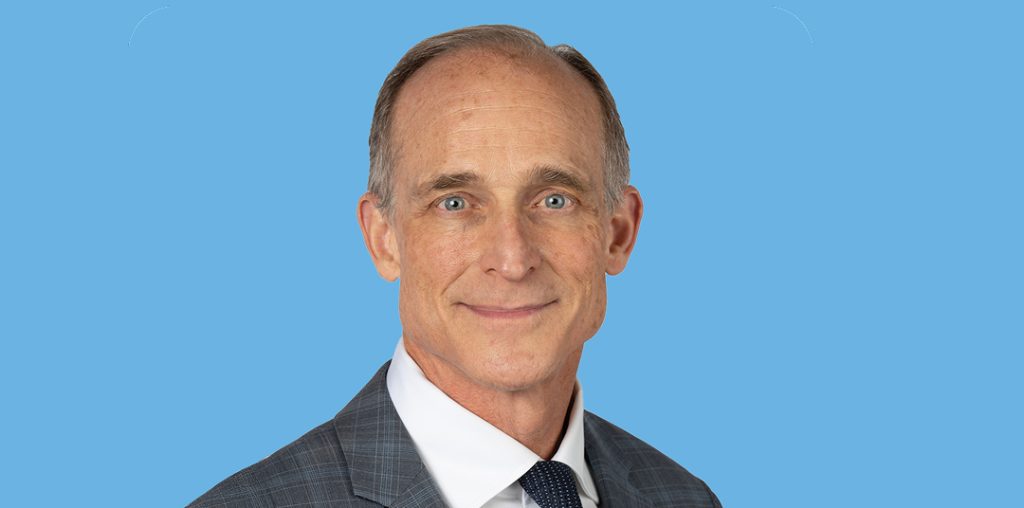 Portrait of a smiling, middle-aged man in a dark gray suit with a white shirt against a blue background.