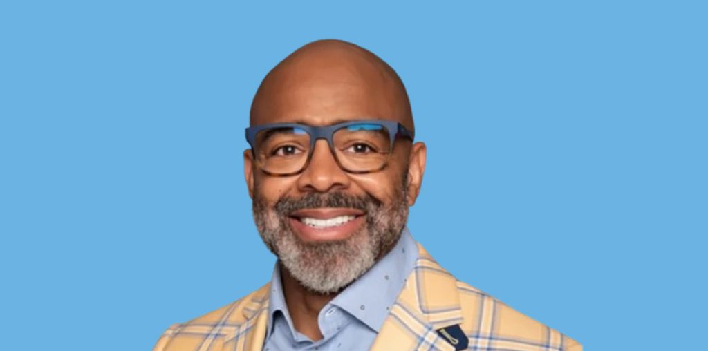 Smiling bald man with a gray beard wearing blue glasses and a yellow plaid blazer against a blue background.