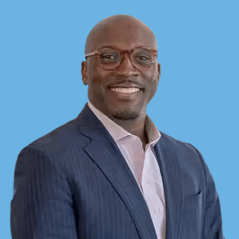 Smiling bald man wearing a navy pinstripe suit, light pink shirt, and glasses, against a blue background.
