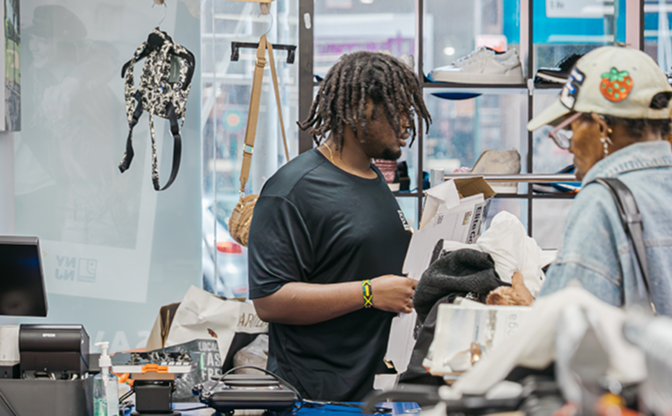 Young shopper with dreadlocks in a black t-shirt browses a box in a sneaker store, with shoe displays behind.