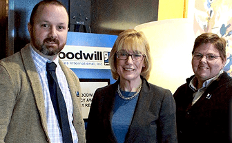 Three business professionals pose for a photo at a Goodwill event, with banners in the background.