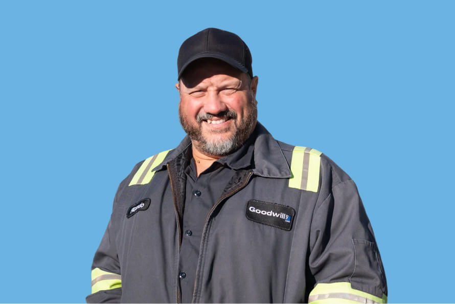 Smiling worker in a gray Goodwill uniform with reflective stripes against a blue background.