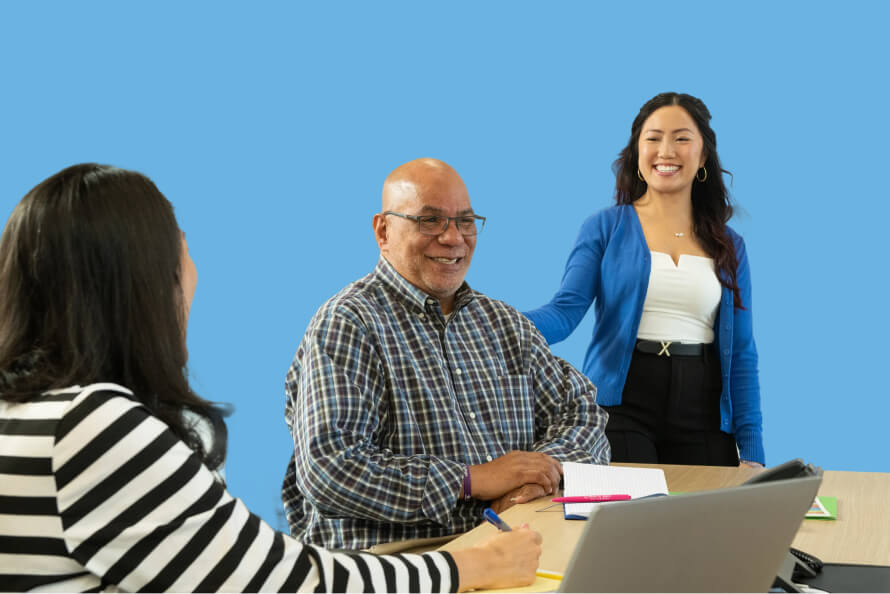 Smiling man in a plaid shirt sits at a table with two coworkers during a meeting, while a woman in blue stands beside him against a blue backdrop.