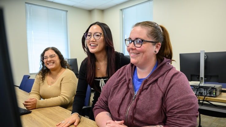 Three women in a computer lab smile while looking at a monitor.