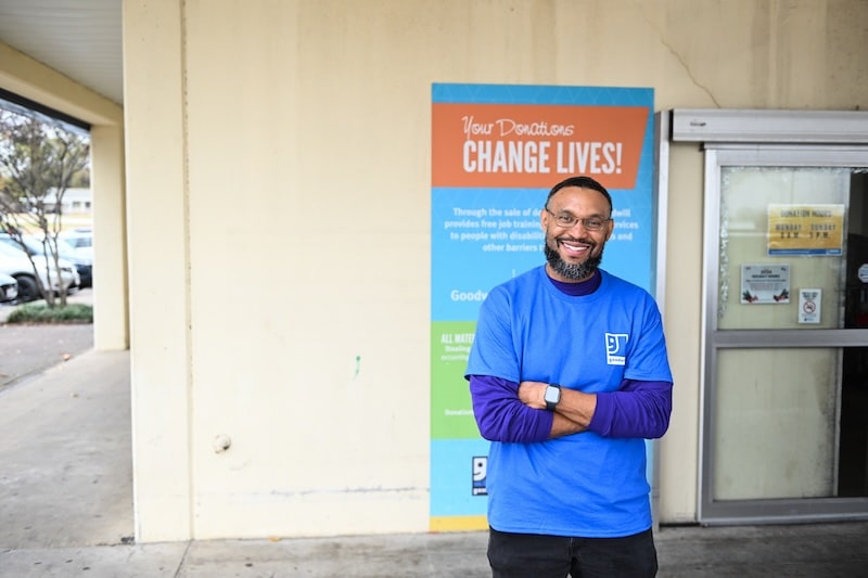 Smiling man in a blue donation-center shirt stands with arms crossed in front of a sign that reads Donations Change Lives.