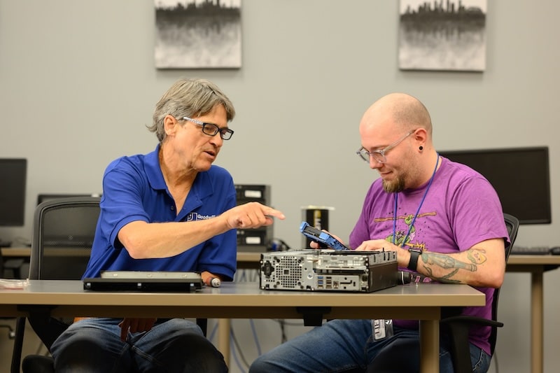 Two men collaborate at a workshop table, one explaining as the other inspects computer hardware.