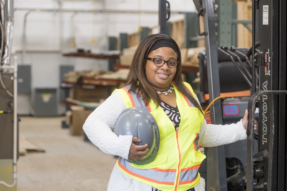 Smiling woman in a yellow safety vest holding a hard hat beside a forklift in a warehouse.