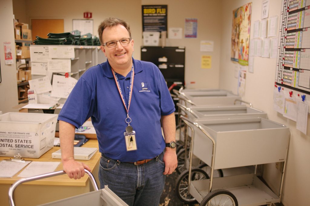 Smiling postal worker in a blue polo shirt leaning on a cart in a busy mailroom.