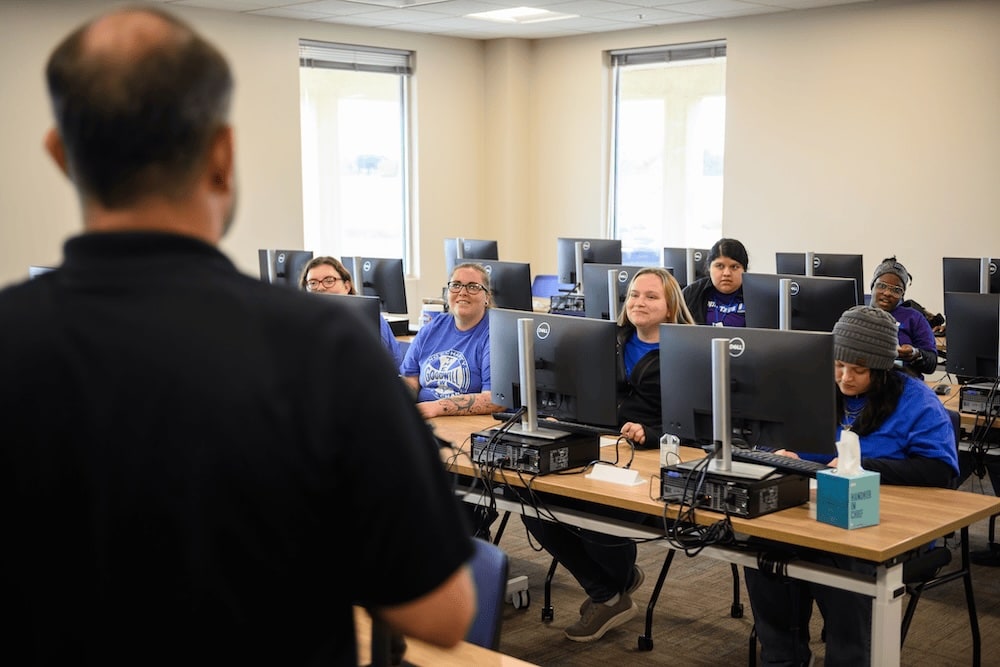 Instructor leads a computer class as students sit at desks with Dell monitors in a bright lab.