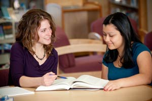 Two women study together at a table with an open notebook in a library.