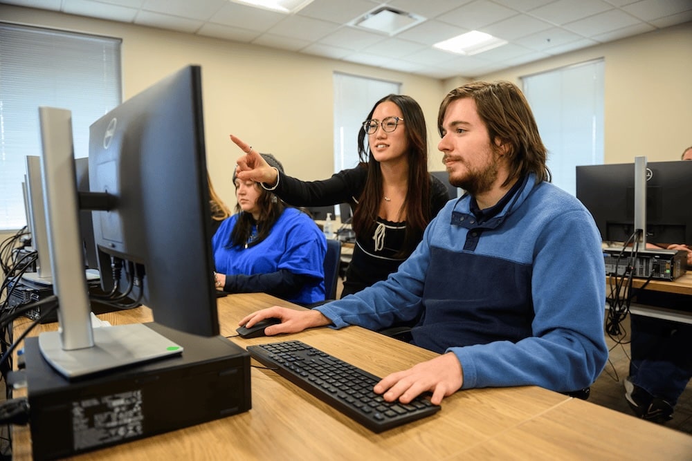 Instructor points at computer screen as a student uses keyboard in a classroom computer lab