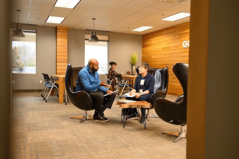 Two professionals discuss at a small wooden table in a modern open office, with black lounge chairs and wooden panel walls.