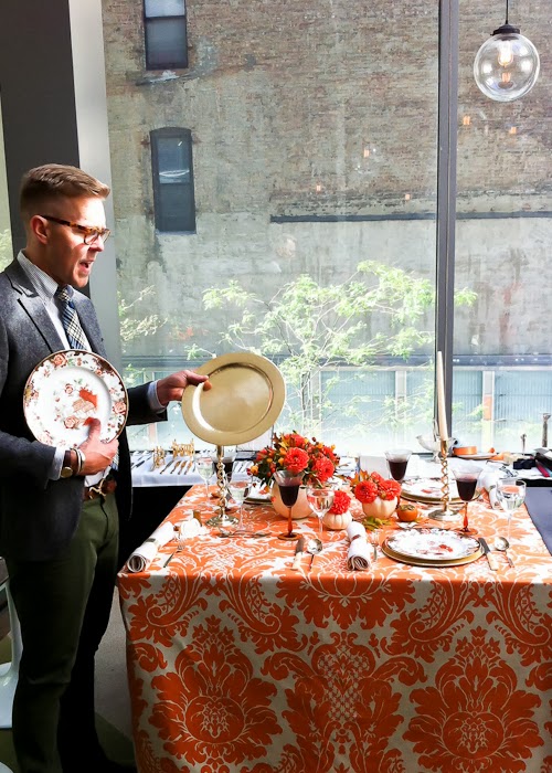 Man in a suit arranges a formal dining table with orange damask cloth, floral centerpiece, and place settings by a window.