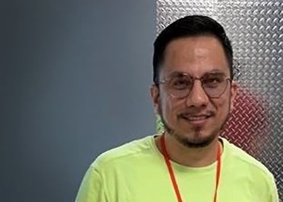 Portrait of a man wearing a neon lime shirt and red lanyard, glasses, against a blue wall and metallic panel.
