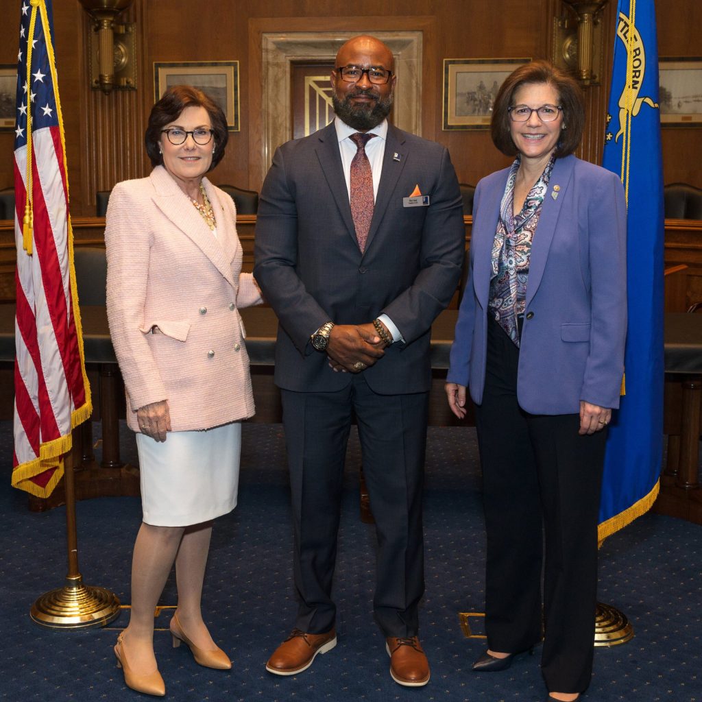 Three officials posing in a government chamber with flags in the background.