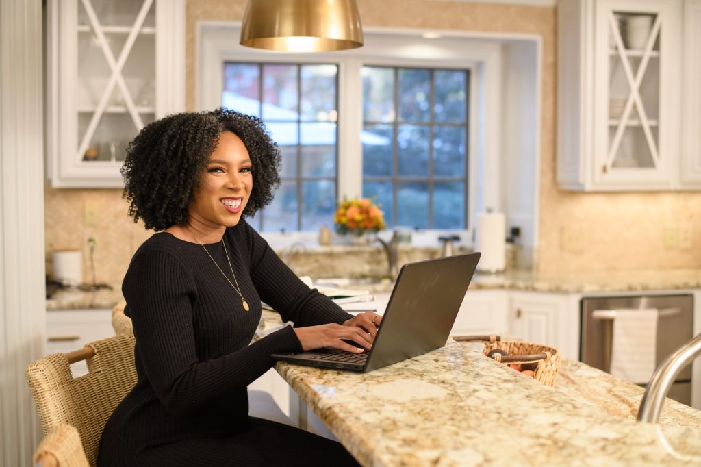 Smiling woman with curly hair typing on a laptop at a granite kitchen island