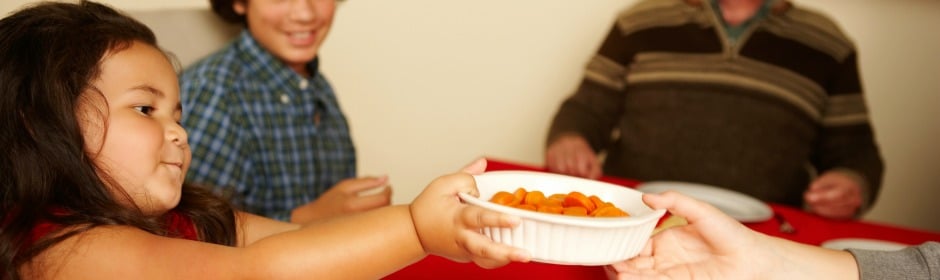 Young girl reaches to receive a white bowl of orange snacks from an adult at a table, with smiling people in the background.