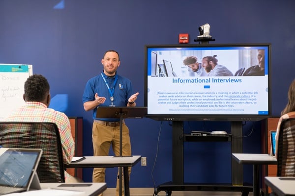 Male presenter in a blue shirt leads a workshop on informational interviews for a small classroom audience, with a large screen displaying the slide.