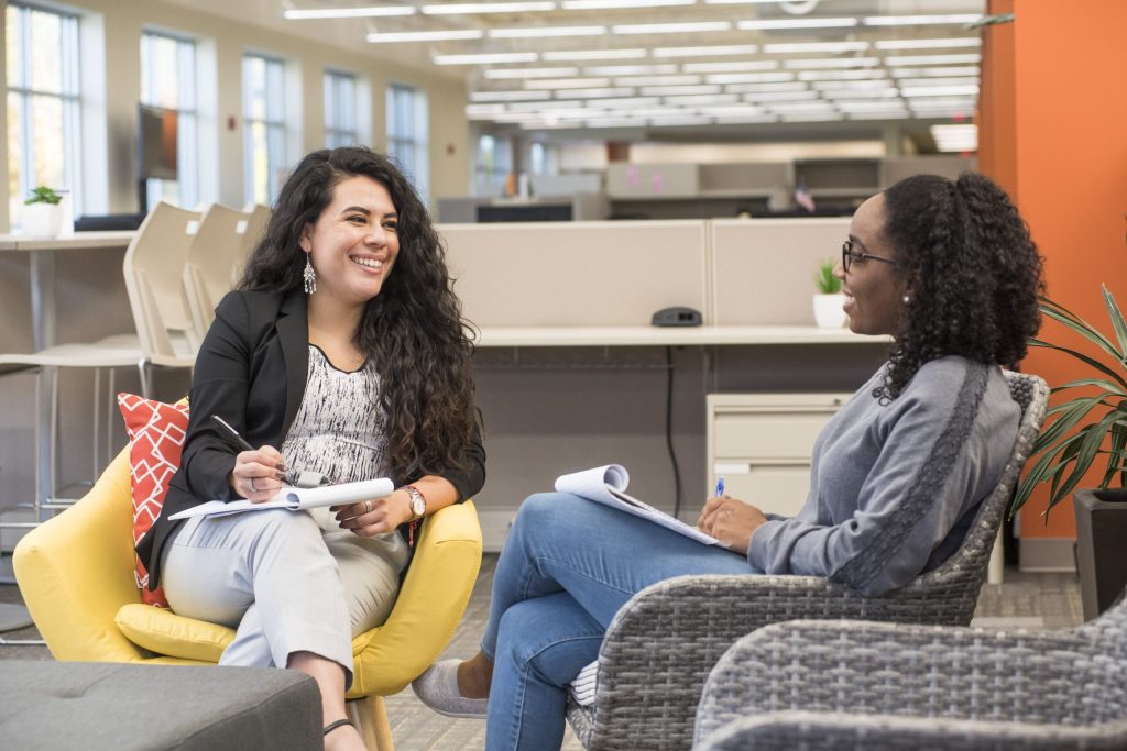 Two professionals in a bright office lounge sit in conversation; one takes notes.