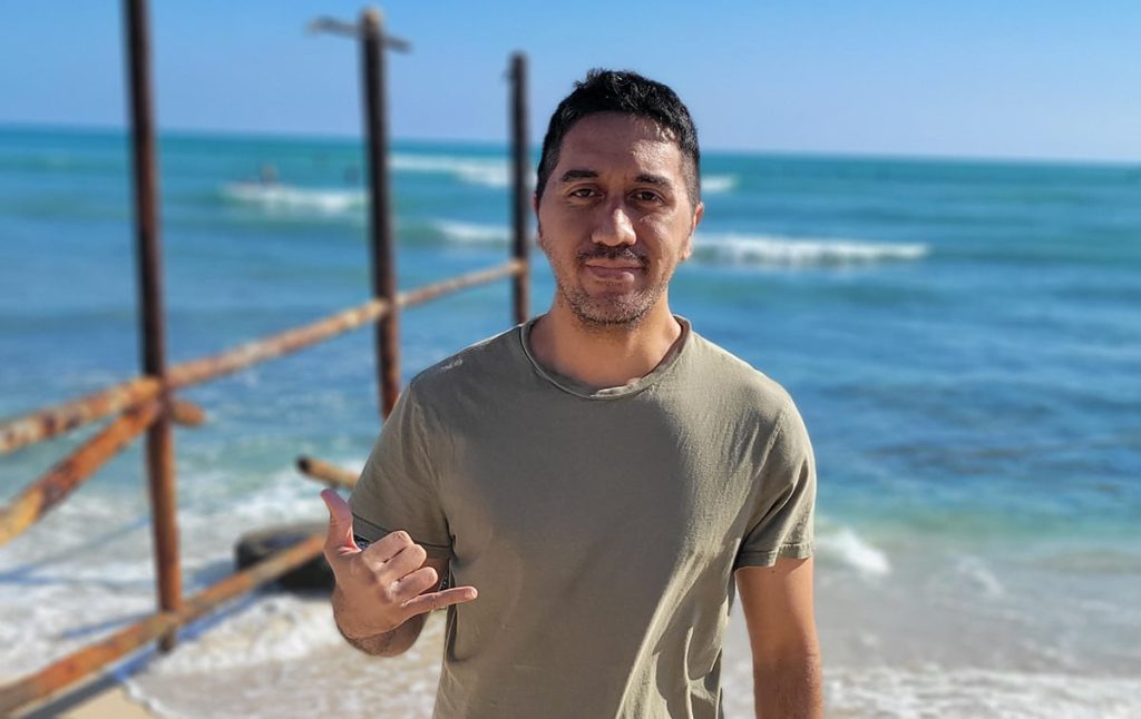 Man at the beach wearing an olive shirt, making a shaka gesture with the ocean behind.