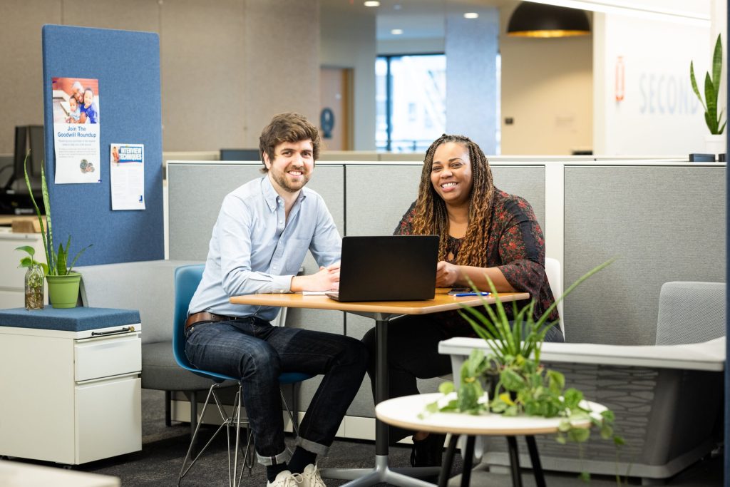 Two coworkers sit at a round table in a bright, modern office, smiling at the camera with a laptop between them.