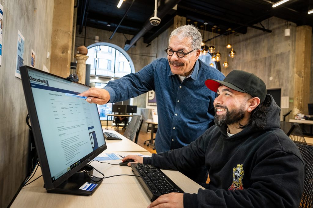 Two men collaborate at a desk with a large monitor showing a form as the older man points at the screen.