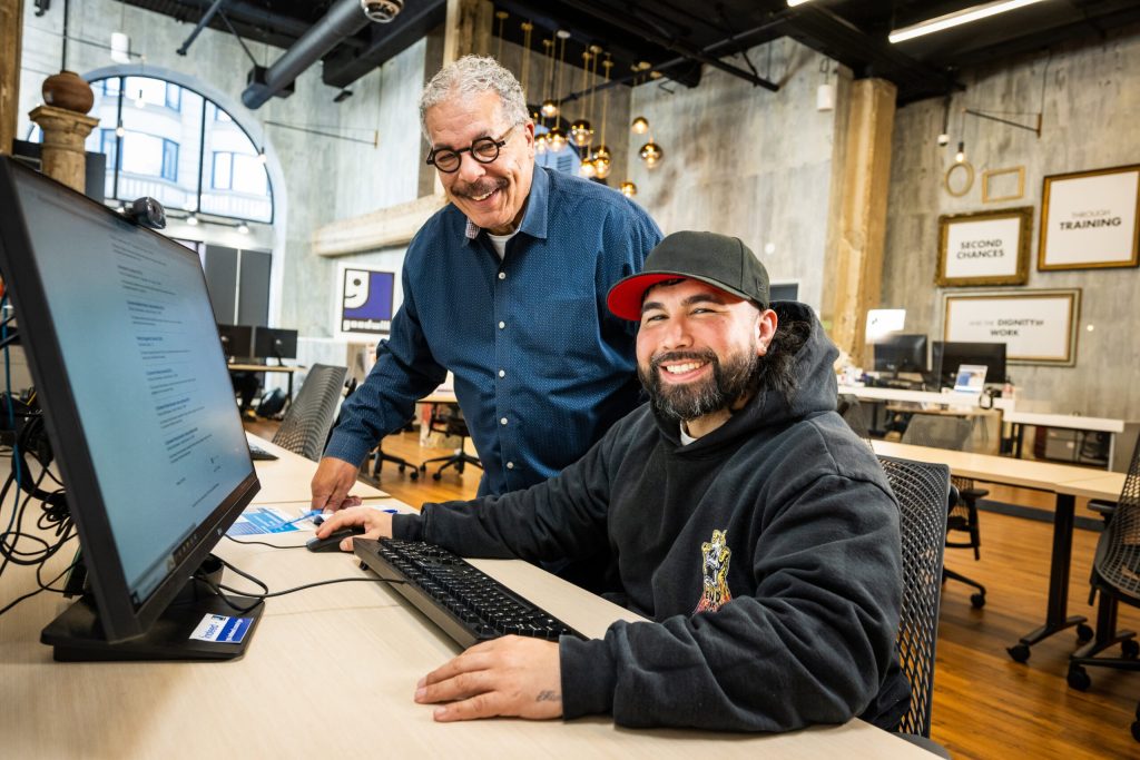 Two men collaborate at a computer in a modern coworking space; one sits at a desk while the other stands nearby.
