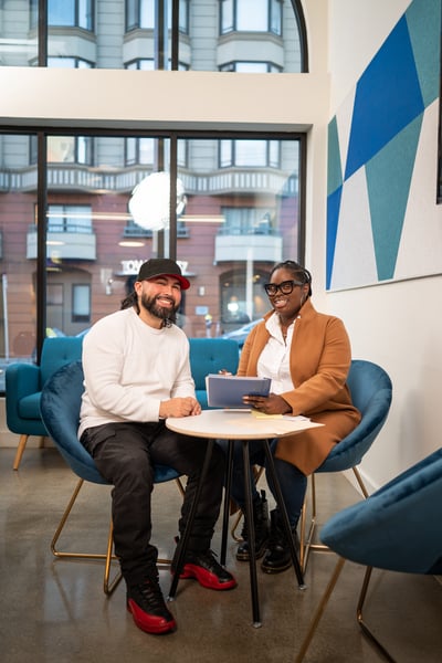 Two colleagues seated at a round table in a modern lounge, smiling at the camera with a tablet on the table.