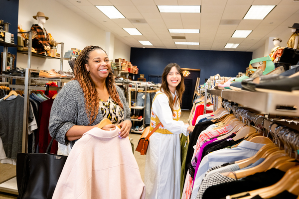 Two women shopping in a bright clothing store, smiling while holding garments.
