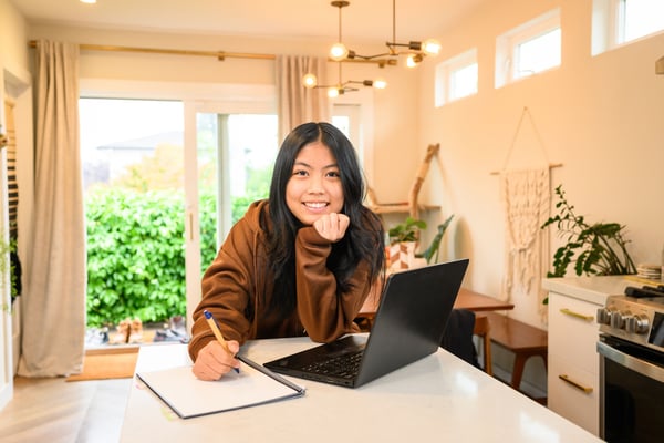 Young woman in brown sweater working at a kitchen island with laptop and notebook.