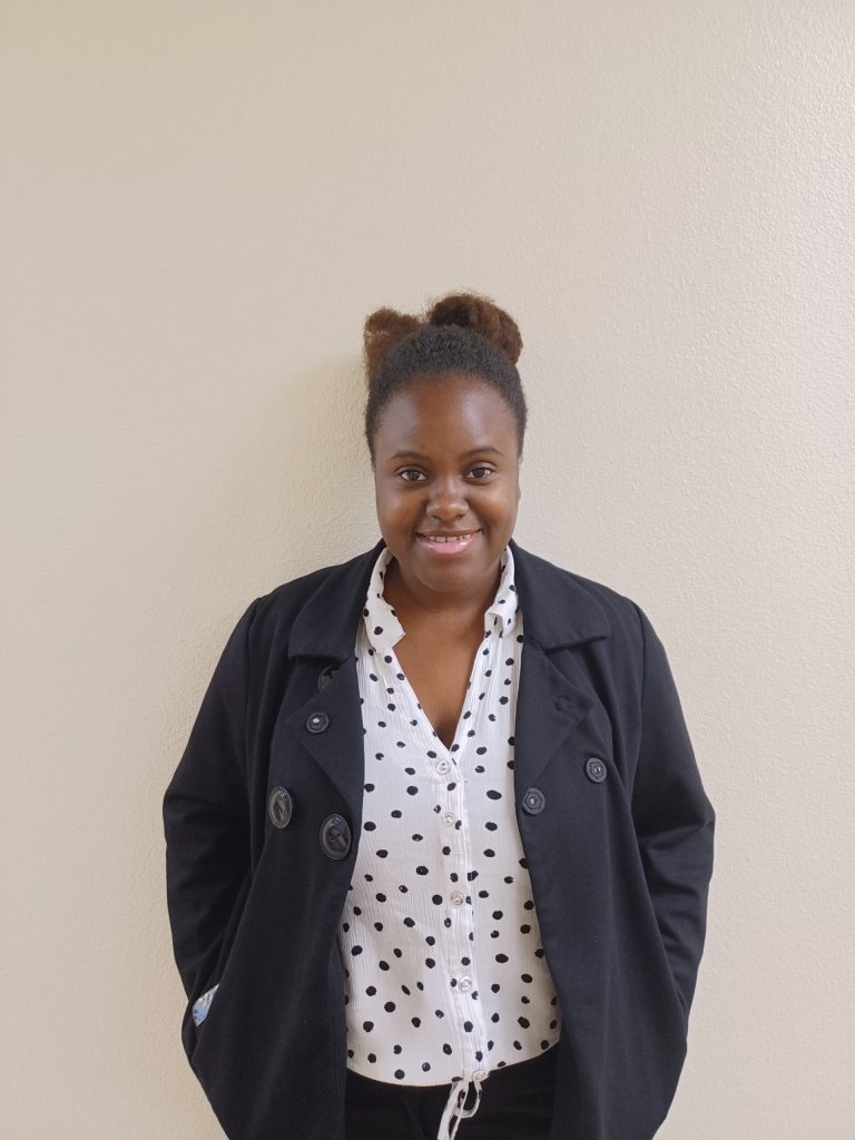 Smiling woman with a dark coat and white polka-dot blouse against a beige wall.
