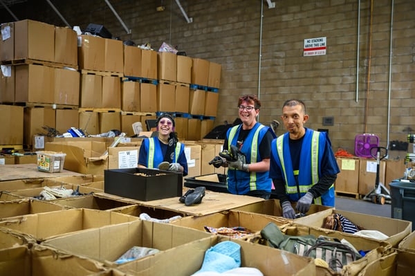 Three volunteers in high-visibility vests sorting donated items in a large warehouse filled with boxes.