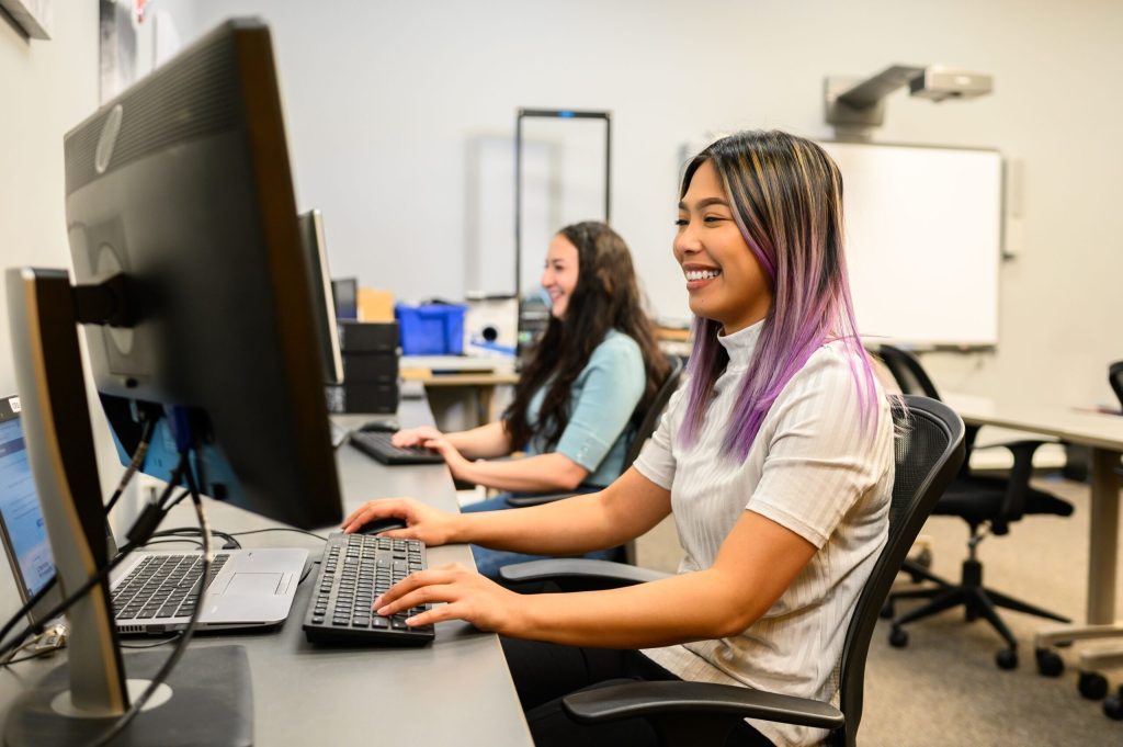 Two women sit at desks using desktop computers in a modern office.
