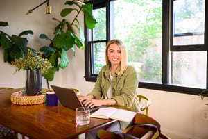 Smiling woman at a wooden table with laptop, notebook, and plants in a bright home office.