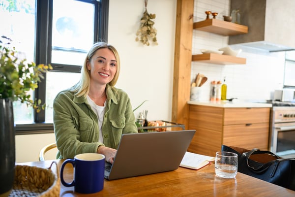 Smiling woman seated at a wooden kitchen table with a laptop, in a bright modern kitchen.