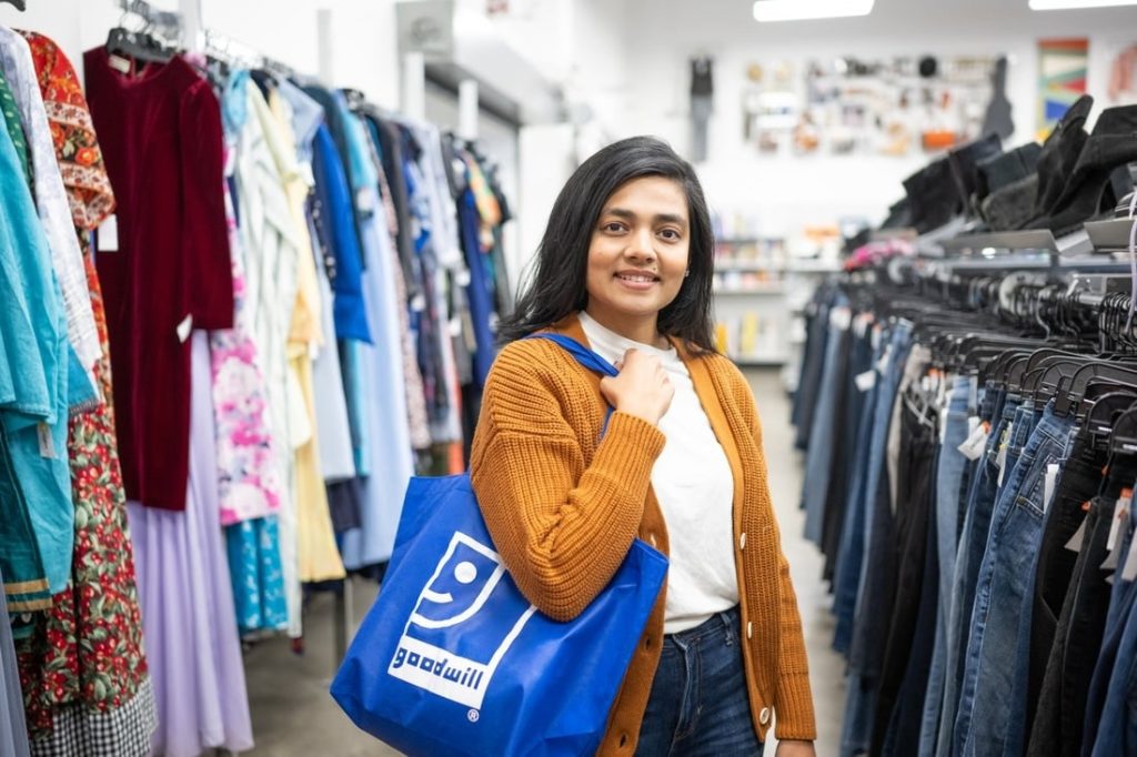 Smiling woman in a thrift store carries a blue Goodwill tote bag, standing between clothing racks.
