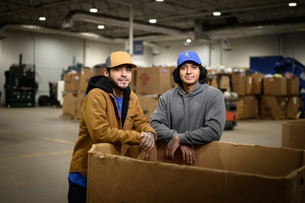 Two workers lean on a large cardboard box in a warehouse, with stacked boxes and industrial shelving in the background.