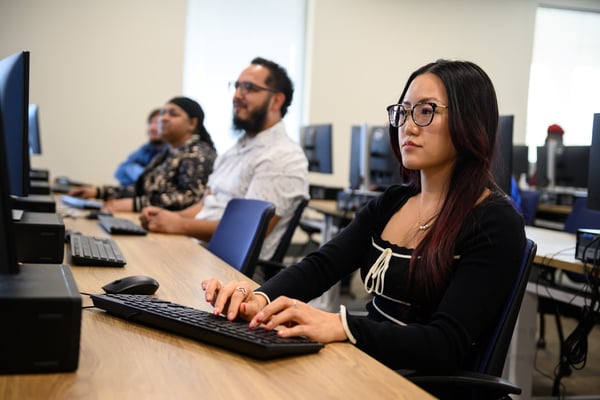 Student with glasses typing at a desktop in a classroom computer lab, with peers at desks in the background.