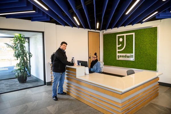 Customer at a Goodwill reception desk in a bright lobby with a moss wall logo and blue ceiling panels.